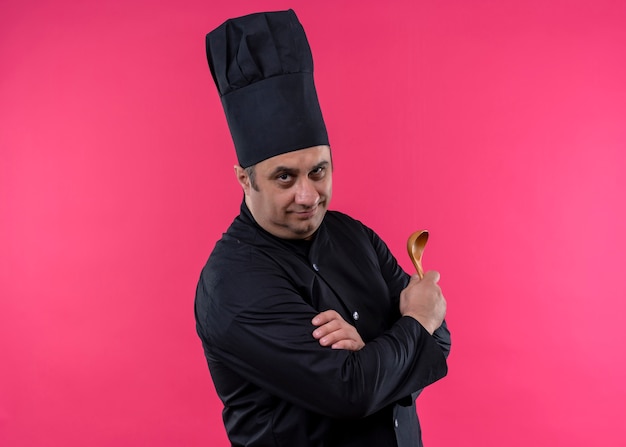 Photo gratuite chef cuisinier mâle vêtu d'un uniforme noir et chapeau de cuisinier tenant une cuillère en bois regardant la caméra avec une expression confiante debout sur fond rose