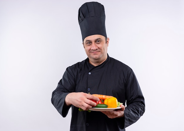 Chef cuisinier mâle vêtu d'un uniforme noir et chapeau de cuisinier tenant la plaque avec des légumes frais regardant la caméra avec le sourire sur le visage debout sur fond blanc