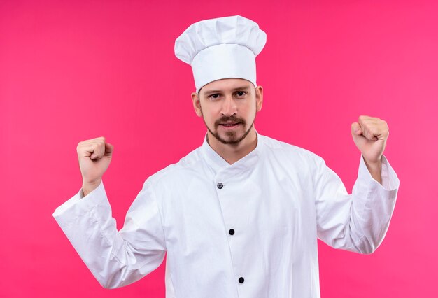 Chef cuisinier professionnel masculin en uniforme blanc et chapeau de cuisinier serrant les poings heureux et excité se réjouissant de son succès debout sur fond rose