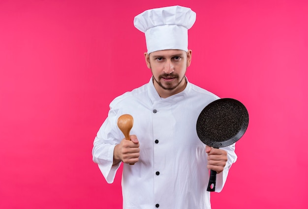 Chef cuisinier professionnel en uniforme blanc et chapeau de cuisinier tenant une casserole et une cuillère en bois regardant la caméra avec un visage sérieux debout sur fond rose
