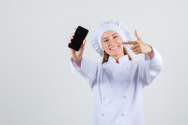 Chef féminin pointant le doigt sur le smartphone en uniforme blanc et à la recherche de bonne humeur. vue de face.