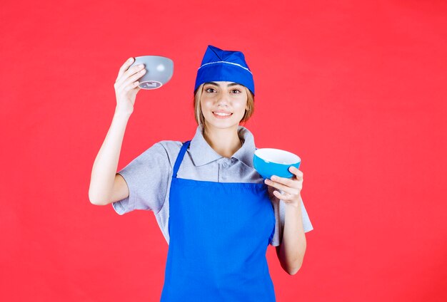 Chef féminin en tablier bleu tenant deux tasses de nouilles en céramique et se confondre au sujet de faire un choix