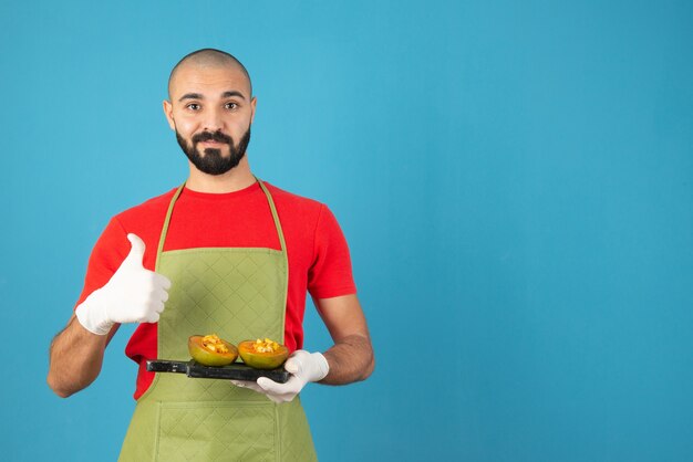 Chef masculin barbu en tablier et gants tenant une planche de bois sombre avec de la pâtisserie.