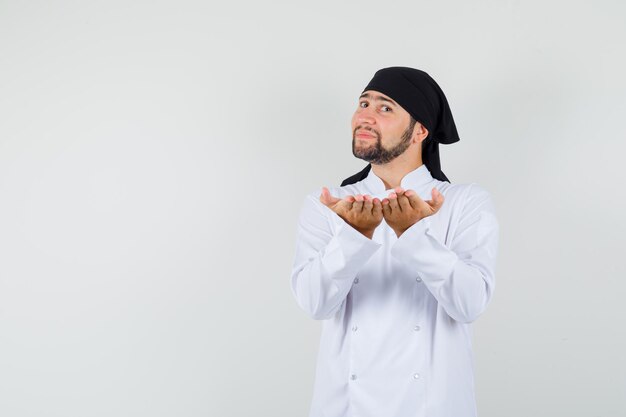 Un chef masculin levant les mains ouvertes comme s'il tenait quelque chose en uniforme blanc et avait l'air joyeux, vue de face.