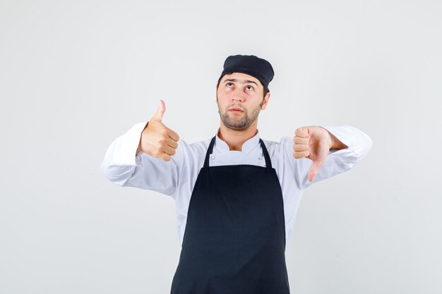 Chef masculin montrant les pouces vers le bas et vers le haut en uniforme, tablier et à la pensif. vue de face.