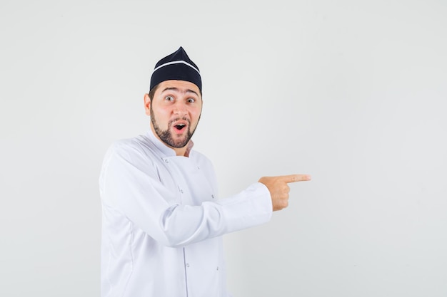 Chef masculin pointant sur le côté en uniforme blanc et semblant étonné. vue de face.