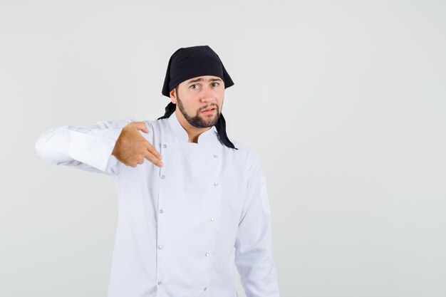 Chef masculin pointant deux doigts sur lui-même en uniforme blanc et l'air perplexe. vue de face.