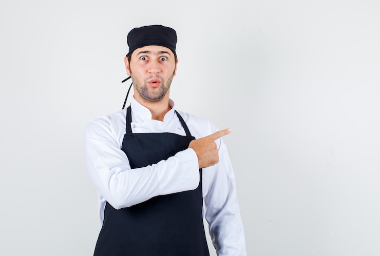 Photo gratuit chef masculin pointant du doigt sur le côté en uniforme, tablier et à la surprise. vue de face.