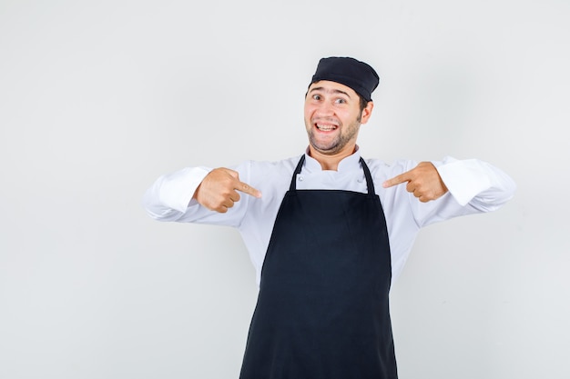 Chef Masculin Pointant Du Doigt Sur Lui-même En Uniforme, Tablier Et à La Vue De Face, Confiant.