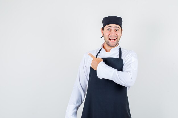 Chef masculin pointant vers l'arrière en uniforme, tablier et à la recherche de bonne humeur. vue de face.