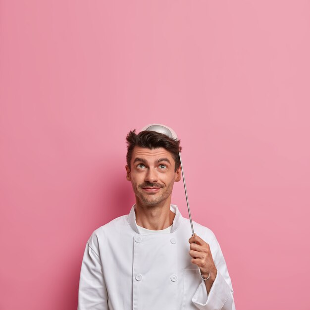 Le chef masculin positif pose avec une louche sur la tête, va préparer la soupe, porte un uniforme blanc, détient des ustensiles de cuisine, fait de la cuisine de restaurant, regarde au-dessus