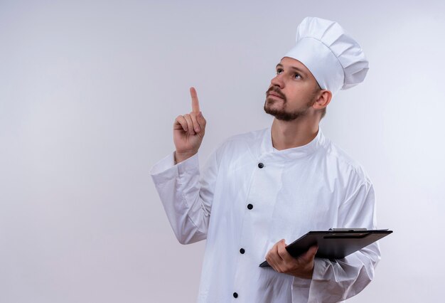 Chef masculin professionnel cuisinier en uniforme blanc et chapeau de cuisinier tenant le presse-papiers avec des pages vierges pointant le doigt vers le haut de se souvenir de chose importante debout sur fond blanc