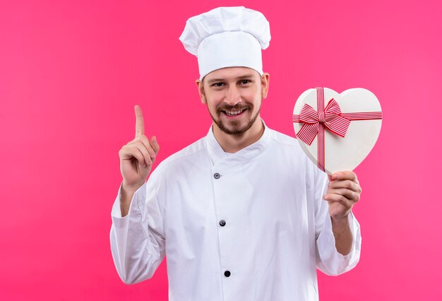 Chef masculin professionnel cuisinier en uniforme blanc et cook hat holding gift box pointer le doigt vers le haut souriant joyeusement sur fond rose