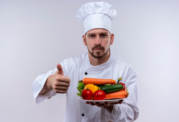 Chef masculin professionnel cuisinier en uniforme blanc et cook hat holding plaque avec des légumes montrant les pouces vers le haut souriant debout sur fond blanc