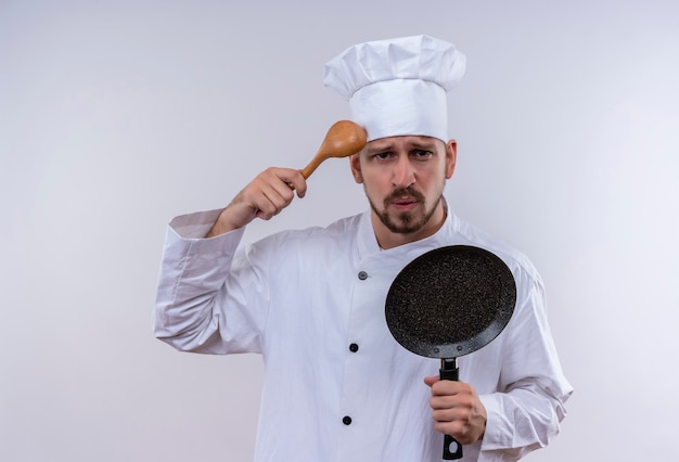 Photo gratuite chef masculin professionnel cuisinier en uniforme blanc et cook hat holding poêle à frire gratter la tête avec une cuillère en bois à confus debout sur fond blanc