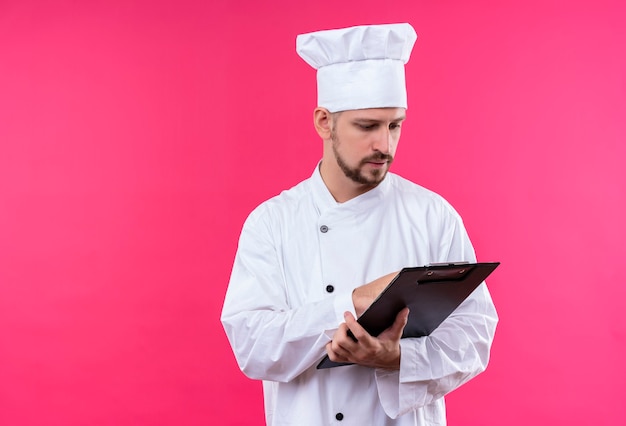 Chef masculin professionnel cuisinier en uniforme blanc et cook hat holding presse-papiers en le regardant avec un visage sérieux debout sur fond rose