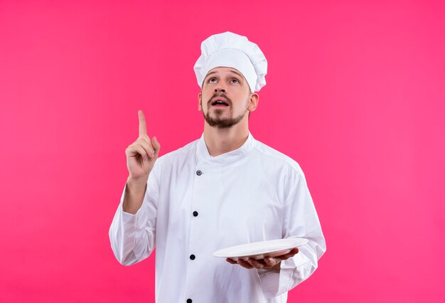 Chef masculin professionnel cuisinier en uniforme blanc et cook hat tenant une assiette emty pointant vers le haut avec le doigt concentré sur une tâche debout sur fond rose