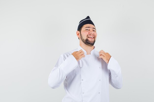 Chef masculin touchant son uniforme avec les mains en uniforme blanc, chapeau et l'air confiant. vue de face.