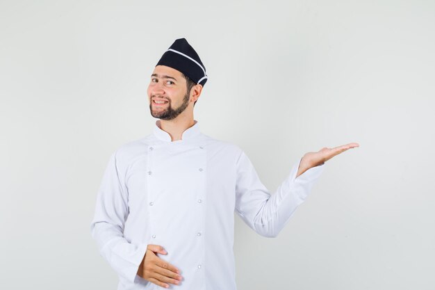 Chef masculin en uniforme blanc levant la main comme montrant quelque chose et ayant l'air heureux, vue de face.