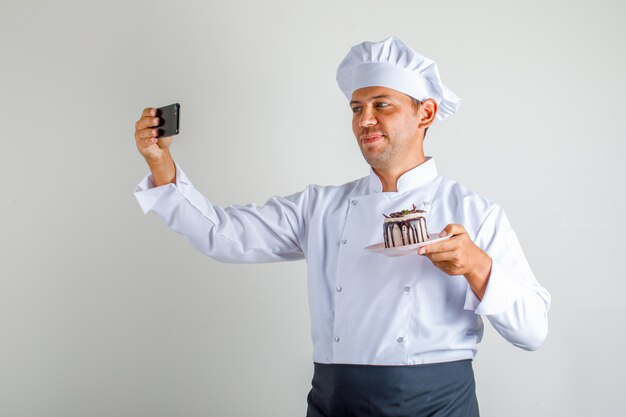 Chef masculin en uniforme, tablier et chapeau prenant selfie avec gâteau au dessert