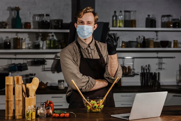 Photo gratuite chef à mi-tir avec masque mélangeant des ingrédients de salade près d'un ordinateur portable