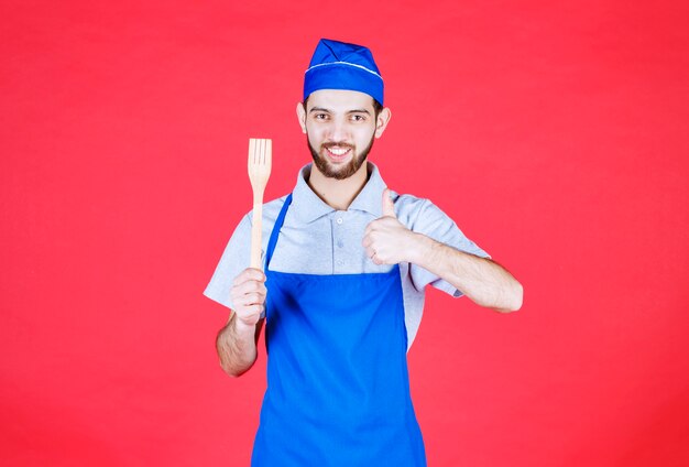 Chef en tablier bleu tenant une spatule en bois et montrant un signe de plaisir.