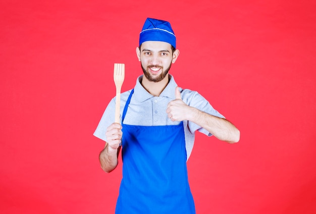 Chef en tablier bleu tenant une spatule en bois et montrant un signe de plaisir.