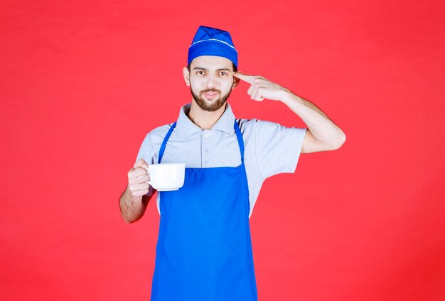 Chef en tablier bleu tenant une tasse en céramique blanche et pensant.