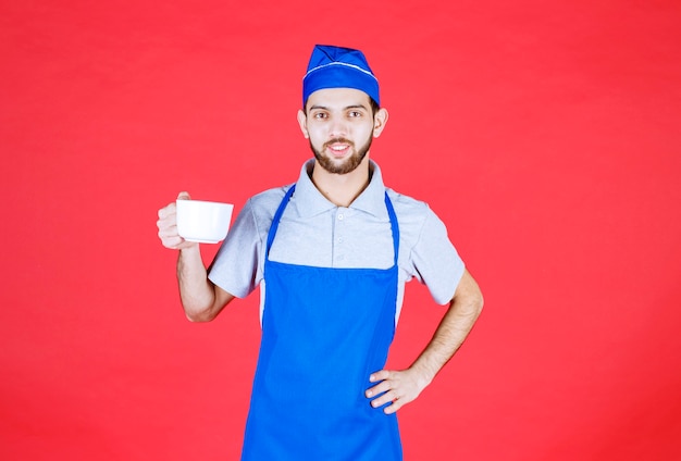 Chef en tablier bleu tenant une tasse en céramique blanche.