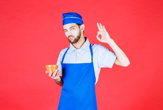 Chef en tablier bleu tenant une tasse en céramique jaune et signe de plaisir showign.