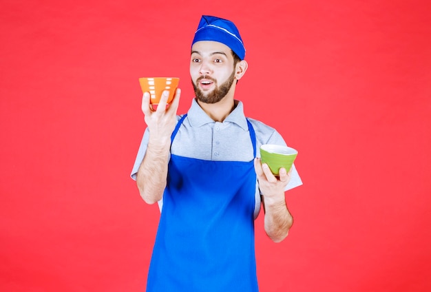 Chef en tablier bleu tenant des tasses en céramique jaune et verte dans les deux mains et a l'air confus.