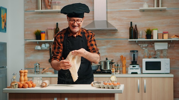 Chef vomir de la pâte pour pizza à la maison dans une cuisine moderne souriant devant la caméra. Habile chef âgé à la retraite portant un uniforme de rotation et de lancer de comptoir à pizza