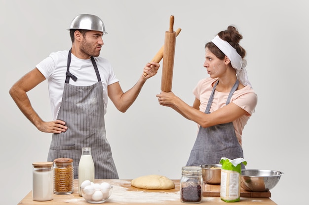 Les Chefs Confiants Ont Une Bataille De Cuisine, Se Regardent Sérieusement, Se Battent Avec Des Rouleaux à Pâtisserie, Se Tiennent Sur Le Côté Près De La Table De La Cuisine Avec De La Pâte Fraîche Et D'autres Ingrédients, Partagent Des Idées Culinaires