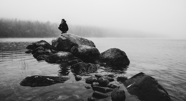 Échelle de gris tourné d'une personne assise sur un gros rocher au milieu de la rivière brumeuse