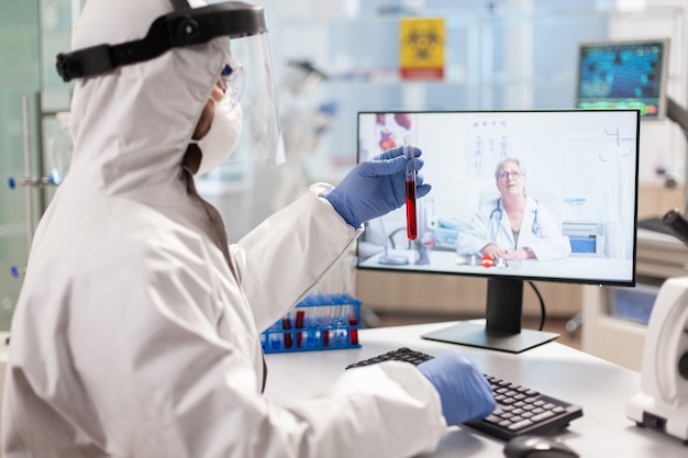 Chercheur de laboratoire discutant avec un appel vidéo vêtu d'un costume en ppe montrant un tube à essai. scientifique montrant un échantillon de sang lors d'une discussion en ligne, recherchant un traitement contre le virus covid19.