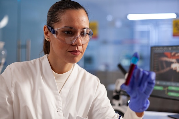 Chercheur scientifique en médecine analysant un échantillon de pot de sang pour un test de développement en laboratoire de chimie. Femme professionnelle avec blouse de laboratoire, lunettes et gants trouvant un traitement pour les soins de santé