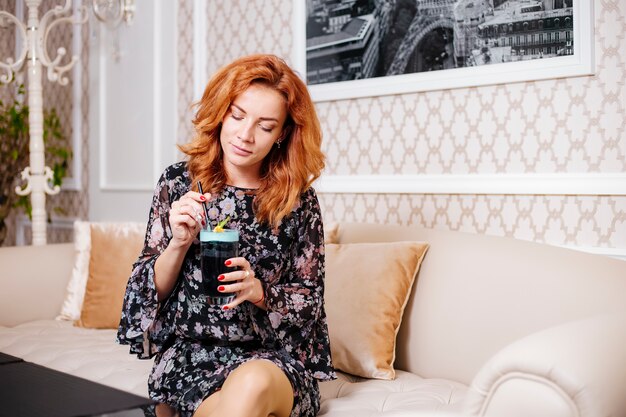 Cheveux roux femme tient un verre à cocktail et est assis dans un canapé de couleur crème