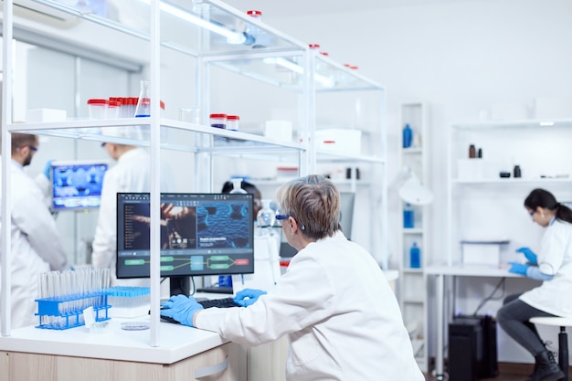 Chimiste médical âgé travaillant sur un ordinateur puissant dans un établissement moderne. scientifique principal dans un laboratoire pharmaceutique effectuant des recherches génétiques portant une blouse de laboratoire avec une équipe en arrière-plan.