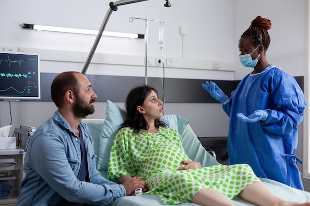 Photo gratuite un chirurgien afro-américain portant des gants de protection et un masque discute avec une femme enceinte expliquant la chirurgie d'accouchement dans le service de l'hôpital.