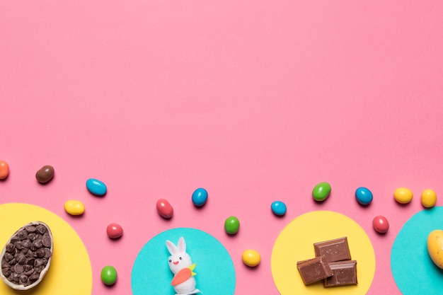 Photo gratuite choco chips; statue de lapin; morceaux de chocolat et bonbons colorés sur fond rose avec espace pour le texte