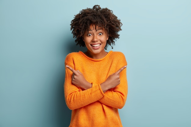 Photo gratuite choisissez une chose. femme optimiste à la peau sombre avec une expression joyeuse, dit que les deux choix sont excellents, croise les mains sur la poitrine, pointe de différents côtés, porte un pull orange, isolé sur un mur bleu