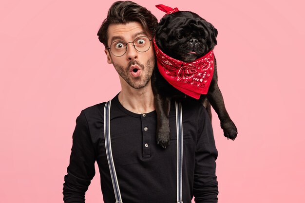 Choqué, beau propriétaire masculin de chien, regarde avec une expression terrible, découvre de mauvaises nouvelles, étant en compagnie d'un animal de compagnie, habillé élégamment, pose ensemble contre un mur rose. Émotions, style de vie