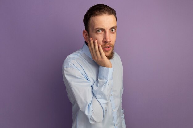 Choqué bel homme blond met la main sur le visage et regarde à l'avant isolé sur mur violet