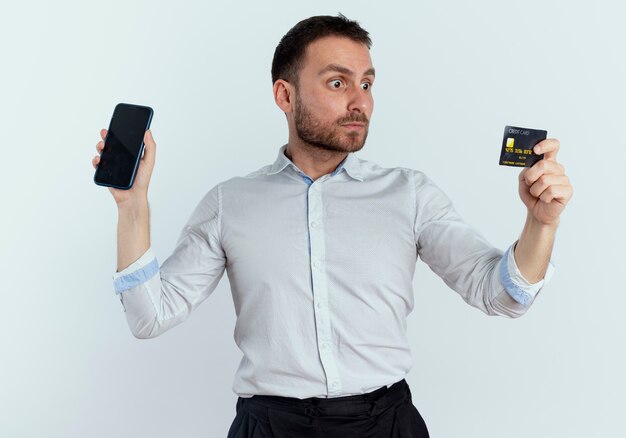 Choqué bel homme tient le téléphone et regarde la carte de crédit isolée sur le mur blanc