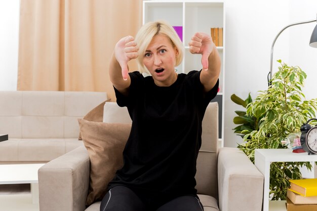 Choqué belle femme russe blonde est assise sur un fauteuil, les pouces vers le bas avec deux mains