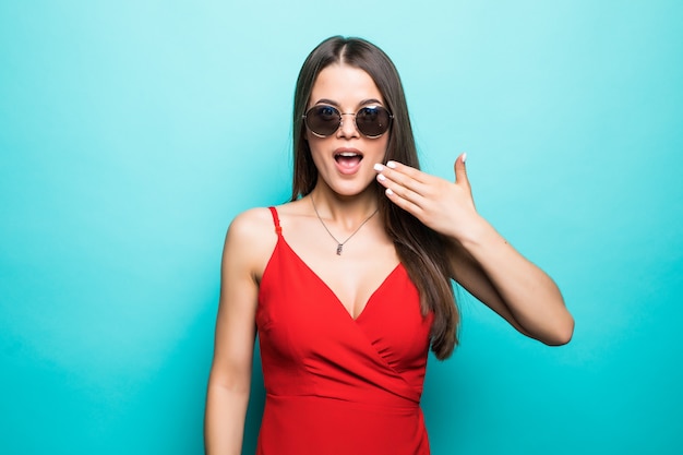 Choqué belle jeune femme en mini robe rouge et lunettes de soleil couvrent la bouche avec la main sur le mur bleu.