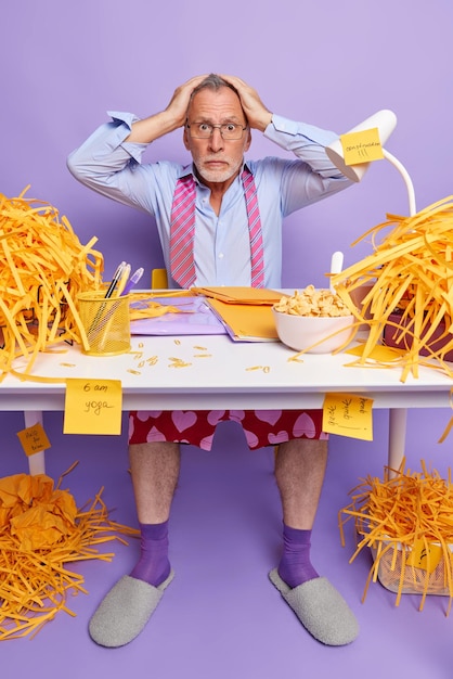 Photo gratuite choqué, embarrassé, un homme aux cheveux gris âgé garde les mains sur la tête pose sur un bureau en désordre ne sait pas par quoi commencer à travailler porte une chemise formelle et une cravate autour du cou travaille à la maison omg concept