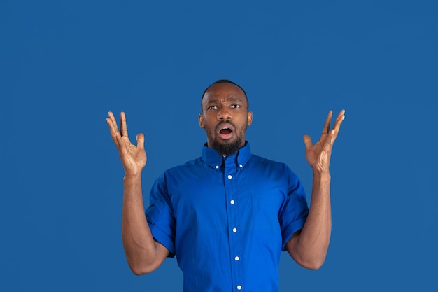 Choqué, étonné. Portrait monochrome de jeune homme afro-américain isolé sur mur bleu. Beau modèle masculin. Émotions humaines, expression faciale, ventes, concept publicitaire. La culture des jeunes.