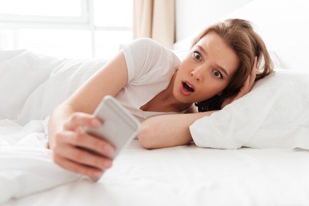 Choqué excité jeune femme au lit, bavardant par téléphone mobile.