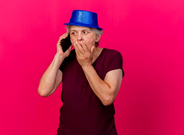 Choqué femme âgée portant chapeau de fête met la main sur la bouche en parlant au téléphone sur rose